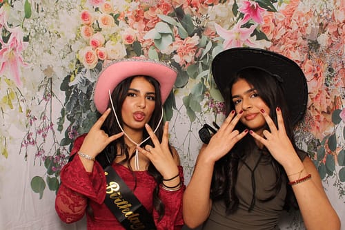Two young women in cowboy hats posing with rock on hand gestures in front of a pink floral wedding backdrop in the West Midlands.