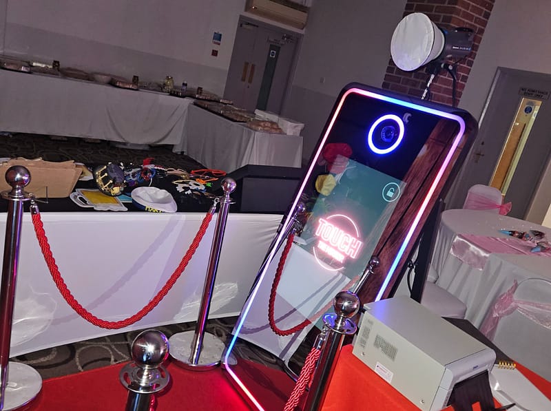 A sleek, smartphone-inspired Magic Mirror photo booth with a glowing purple and red LED border and "Touch the Mirror" prompt, set up on a red carpet in a party venue.