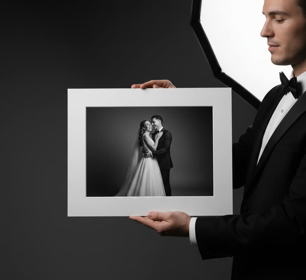 A man in a tuxedo holding a large, white-matted 6x8 black and white wedding portrait print in front of a professional studio softbox light