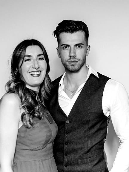 A high-resolution black and white photo booth portrait of a smiling man and woman, featuring crisp studio lighting and a clean white background.