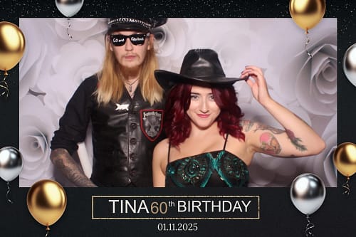 A man with long blonde hair in a leather vest and a woman with red hair in a cowboy hat posing in a photo booth with a 60th birthday digital frame.