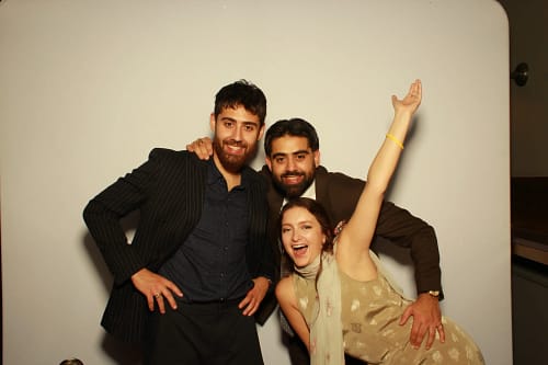 Three wedding guests posing joyfully in a photo booth at Hampton Manor, featuring two men in suits and a woman in a gold dress with her arm raised.