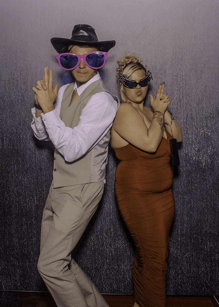 Two wedding guests wearing oversized pink sunglasses and a cowboy hat posing playfully for a photo booth.